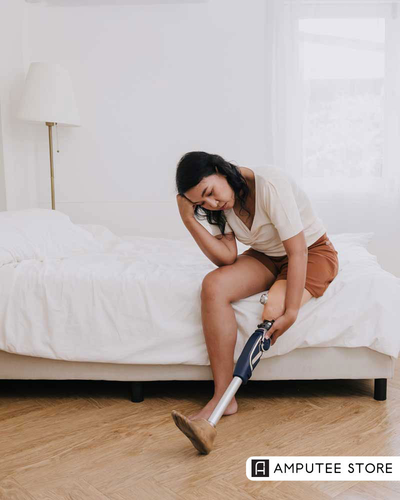 A woman with a prosthetic leg sits on the edge of a bed, gently touching her residual limb with a thoughtful expression.