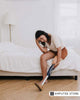 A woman with a prosthetic leg sits on the edge of a bed, gently touching her residual limb with a thoughtful expression.
