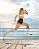 Female below-knee amputee runs outdoors with a blade prosthesis during hot weather beside the water.