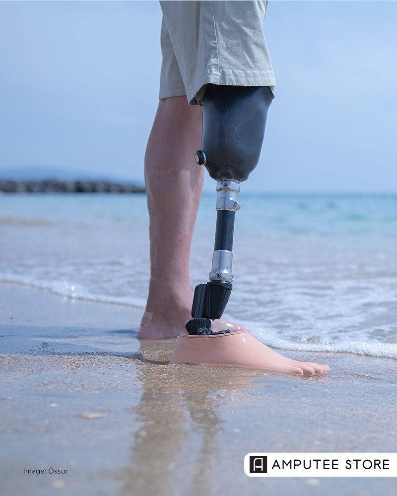 Lower-limb prosthesis user standing on the shoreline as waves wash over the prosthetic foot, highlighting how to care for prosthetic legs around pools, beaches, and showers.