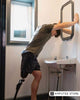 Man with a prosthetic leg leaning over a sink, highlighting daily grooming challenges like shaving for prosthesis users.